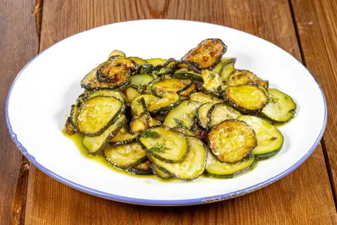 Piatto di zucchine alla poverella, fette sottili e dorate condite con olio e erbe aromatiche, su un piatto bianco con bordo blu.