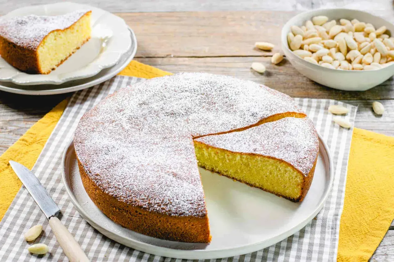 Torta soffice con farina di mandorle, spolverata di zucchero a velo. Accanto, una ciotola di mandorle intere e una fetta già servita.