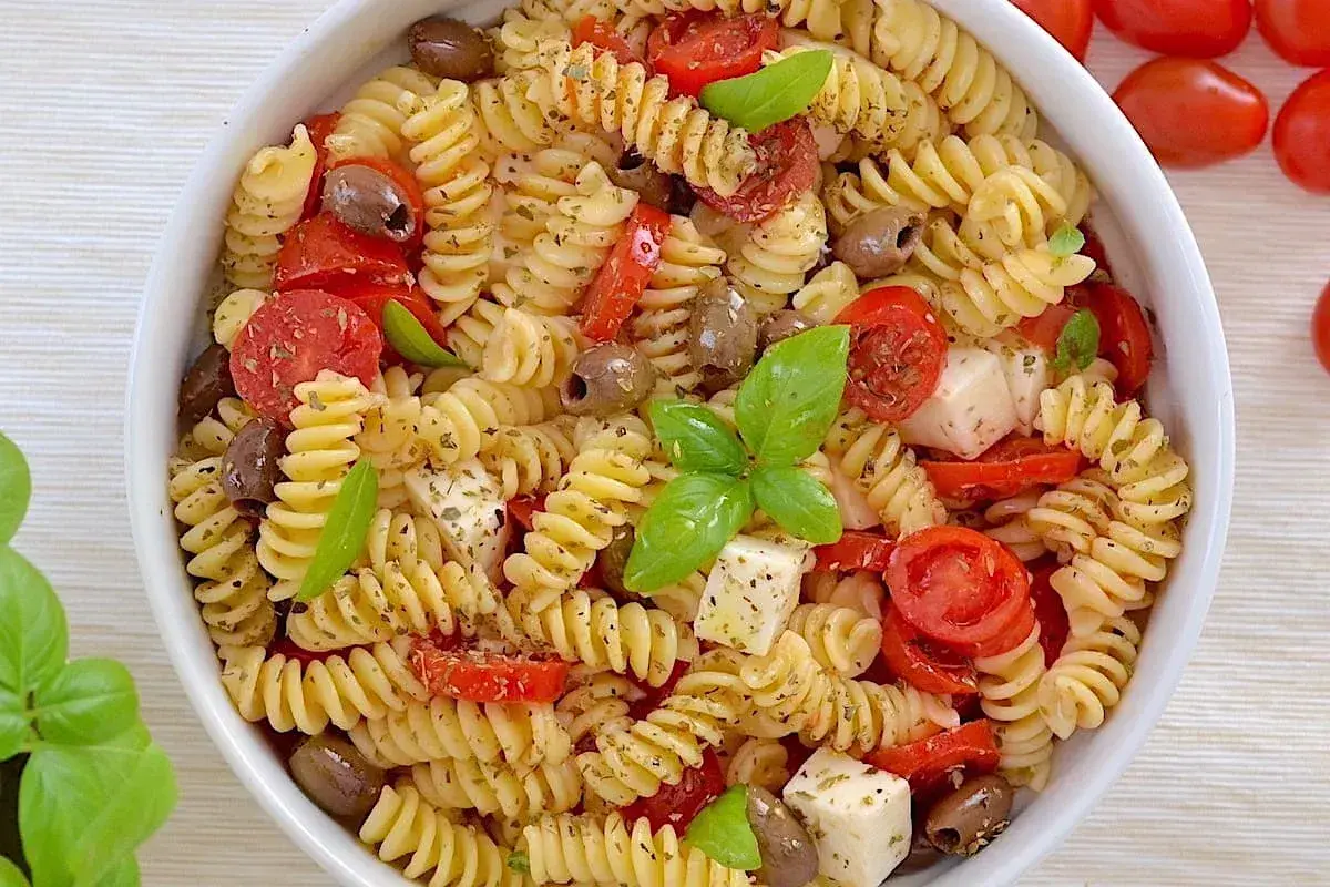 Un piatto invitante di pasta fredda con pomodorini, olive, cubetti di mozzarella e basilico fresco. Perfetta per un'estate gustosa.