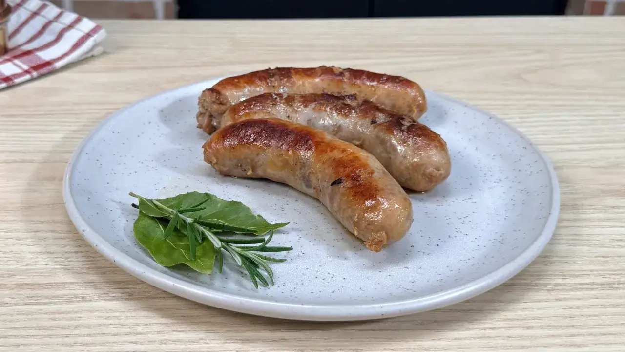 Tre salsicce dorate e succulente, appena cotte in padella, riposano su un piatto con rametti di rosmarino e alloro.