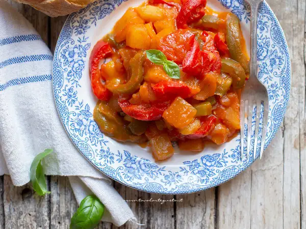 Piatto di peperonata con patate, colorata e saporita, guarnita con basilico fresco su un piatto decorato.