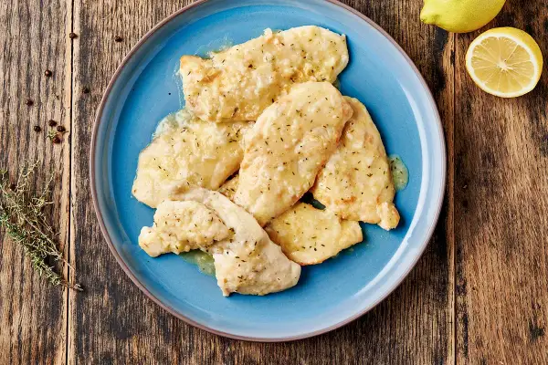 Piatto di pollo al limone su un piatto blu, con limone e rosmarino sullo sfondo.